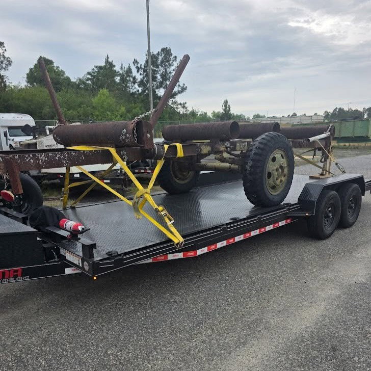 Rusty metal equipment with large wheels strapped onto a flatbed trailer in a parking lot.