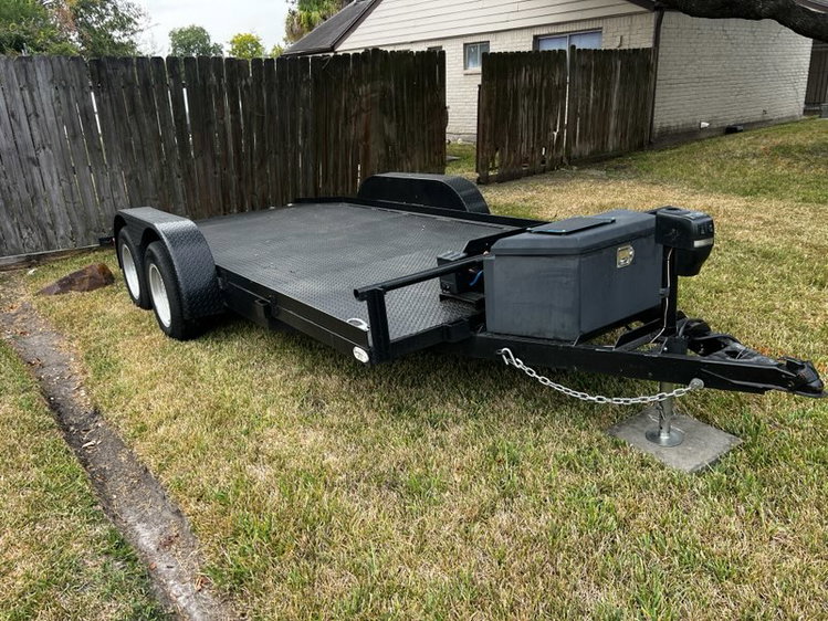 Flatbed utility trailer with dual axles parked on grass near a wooden fence.