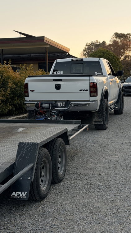 White pickup truck hitched to a flatbed trailer parked on a gravel driveway.