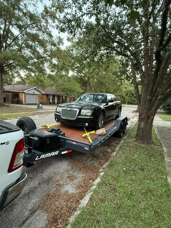 Black sedan strapped onto a black Lamar car hauler trailer with toolbox and spare tire, rented for vehicle transport